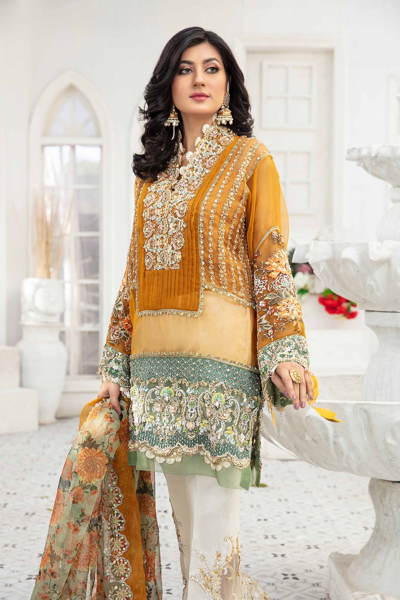 A Pakistani formal dress ensemble featuring a mustard-colored, long flowing gown made of luxurious materials such as silk or chiffon. The gown is adorned with intricate silver work on the neckline, sleeves, and hemline. The model is wearing off-white trousers that complement the gown's color. A printed organza dupatta with delicate floral or geometric designs is draped over the model's head and shoulders, adding a touch of whimsy to the overall look.