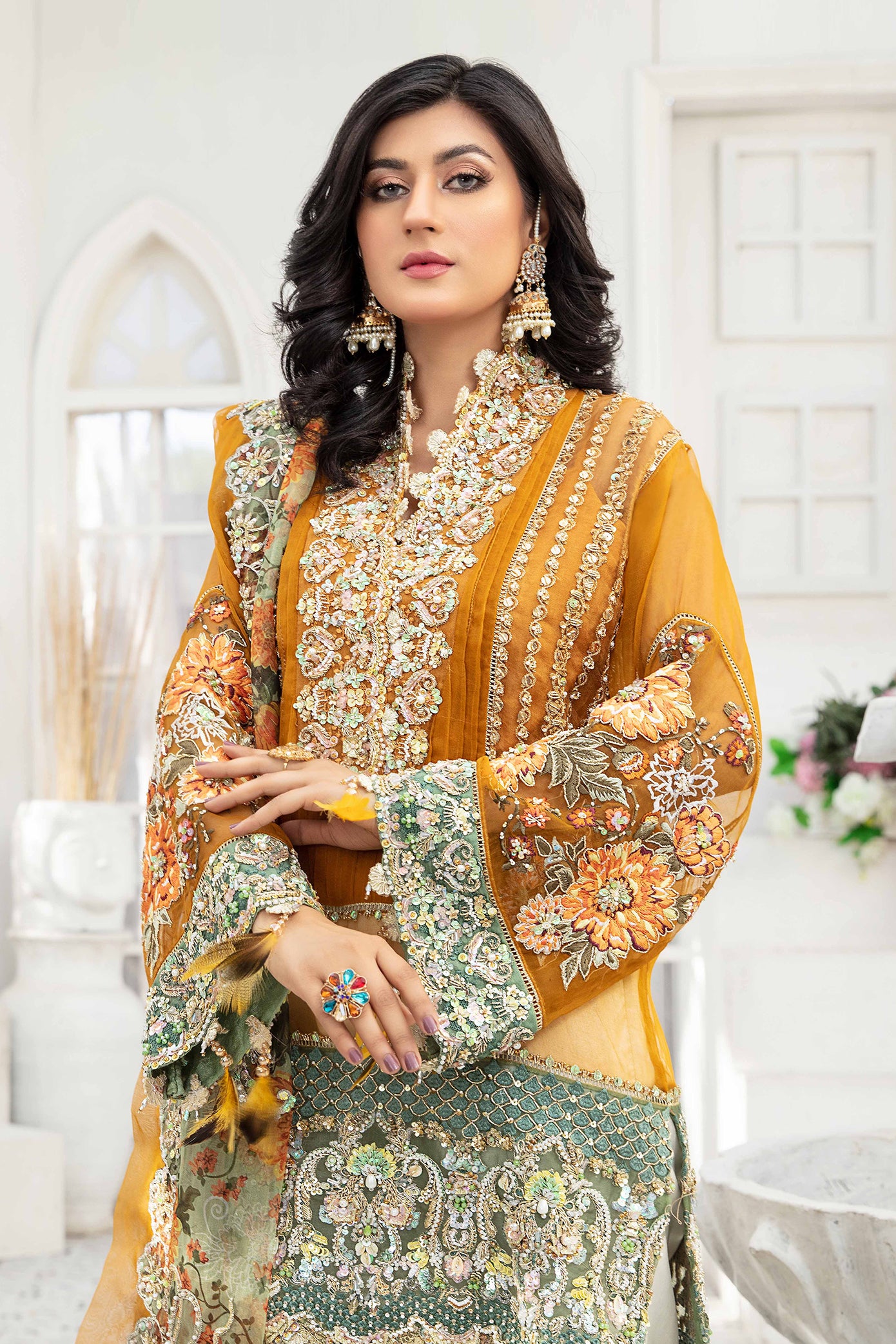 A Pakistani formal dress ensemble featuring a mustard-colored, long flowing gown made of luxurious materials such as silk or chiffon. The gown is adorned with intricate silver work on the neckline, sleeves, and hemline. The model is wearing off-white trousers that complement the gown's color. A printed organza dupatta with delicate floral or geometric designs is draped over the model's head and shoulders, adding a touch of whimsy to the overall look.