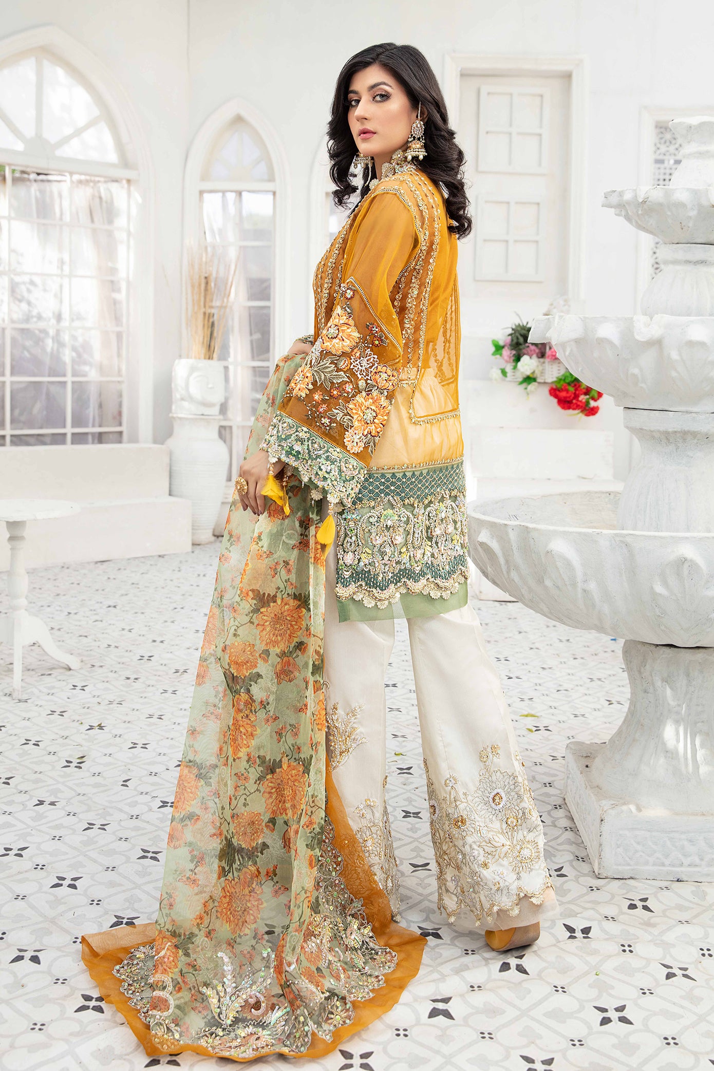 A Pakistani formal dress ensemble featuring a mustard-colored, long flowing gown made of luxurious materials such as silk or chiffon. The gown is adorned with intricate silver work on the neckline, sleeves, and hemline. The model is wearing off-white trousers that complement the gown's color. A printed organza dupatta with delicate floral or geometric designs is draped over the model's head and shoulders, adding a touch of whimsy to the overall look.