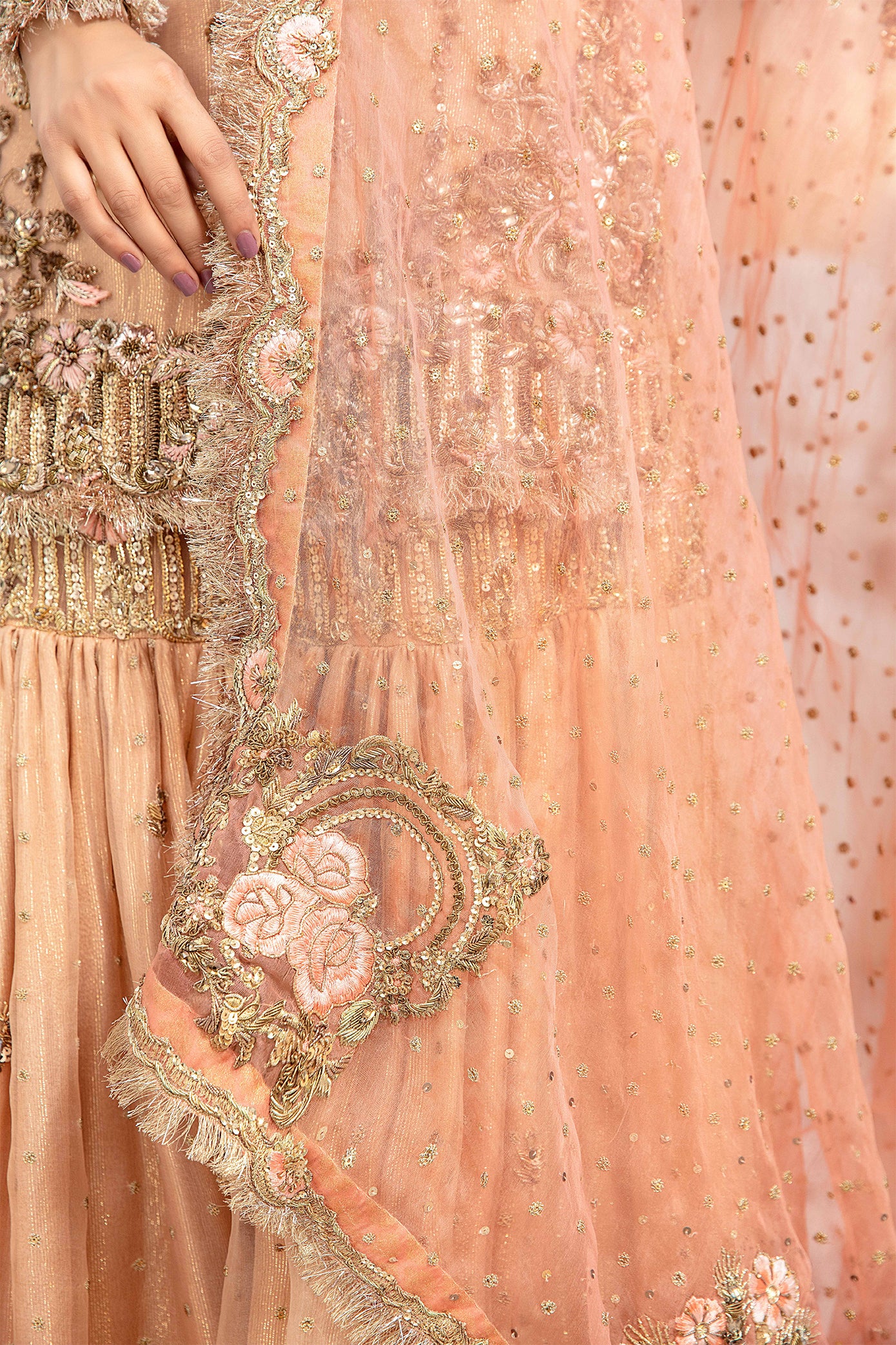 A Peach Gold Pakistani Bridal Dress with intricate gold work on the bodice, sleeves, and hemline. The dress has a fitted silhouette and is made of high-quality fabric that drapes beautifully. The dress is paired with a soft pink chiffon dupatta with intricate gold work on its borders. The bride is standing with a veil draped over her head, looking elegant and regal.