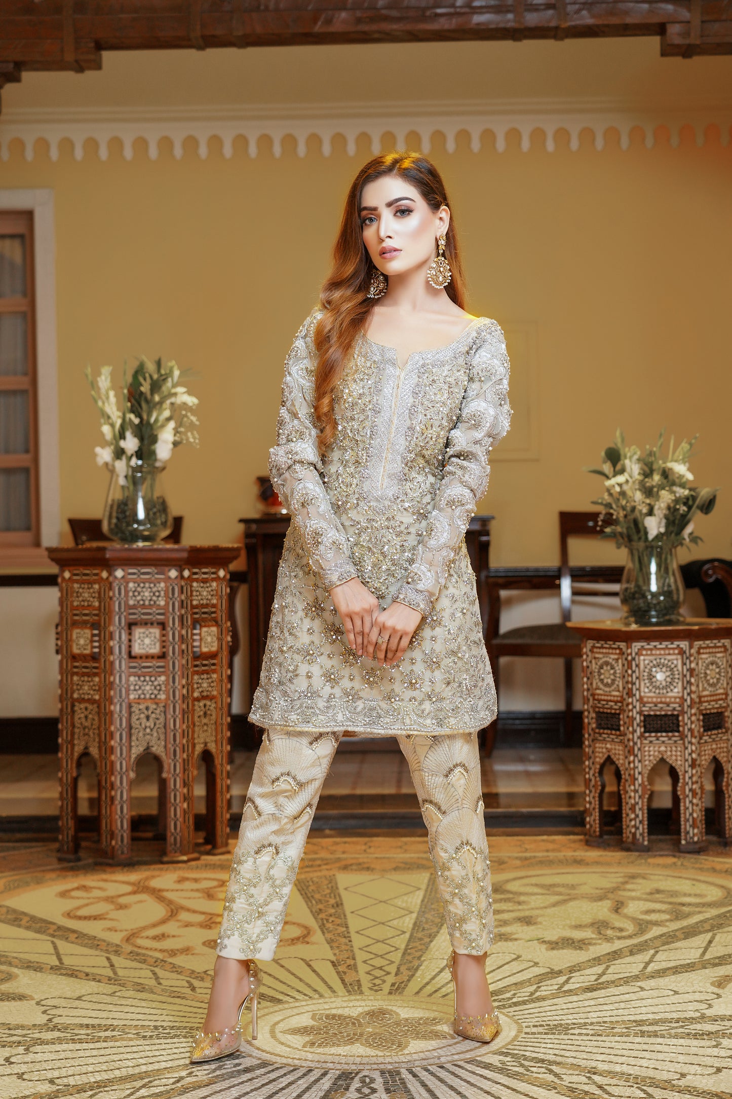 A light gold Pakistani formal dress with gold work and silver work around the neck. The dress has a fitted bodice, high neckline, and long sleeves, with intricate gold work detailing throughout the fabric. Silver work is present around the neckline, creating a beautiful frame for the wearer's face. The flowing skirt adds elegance and grace to the overall look. This dress is perfect for formal occasions and showcases the opulence and refinement of Pakistani fashion.