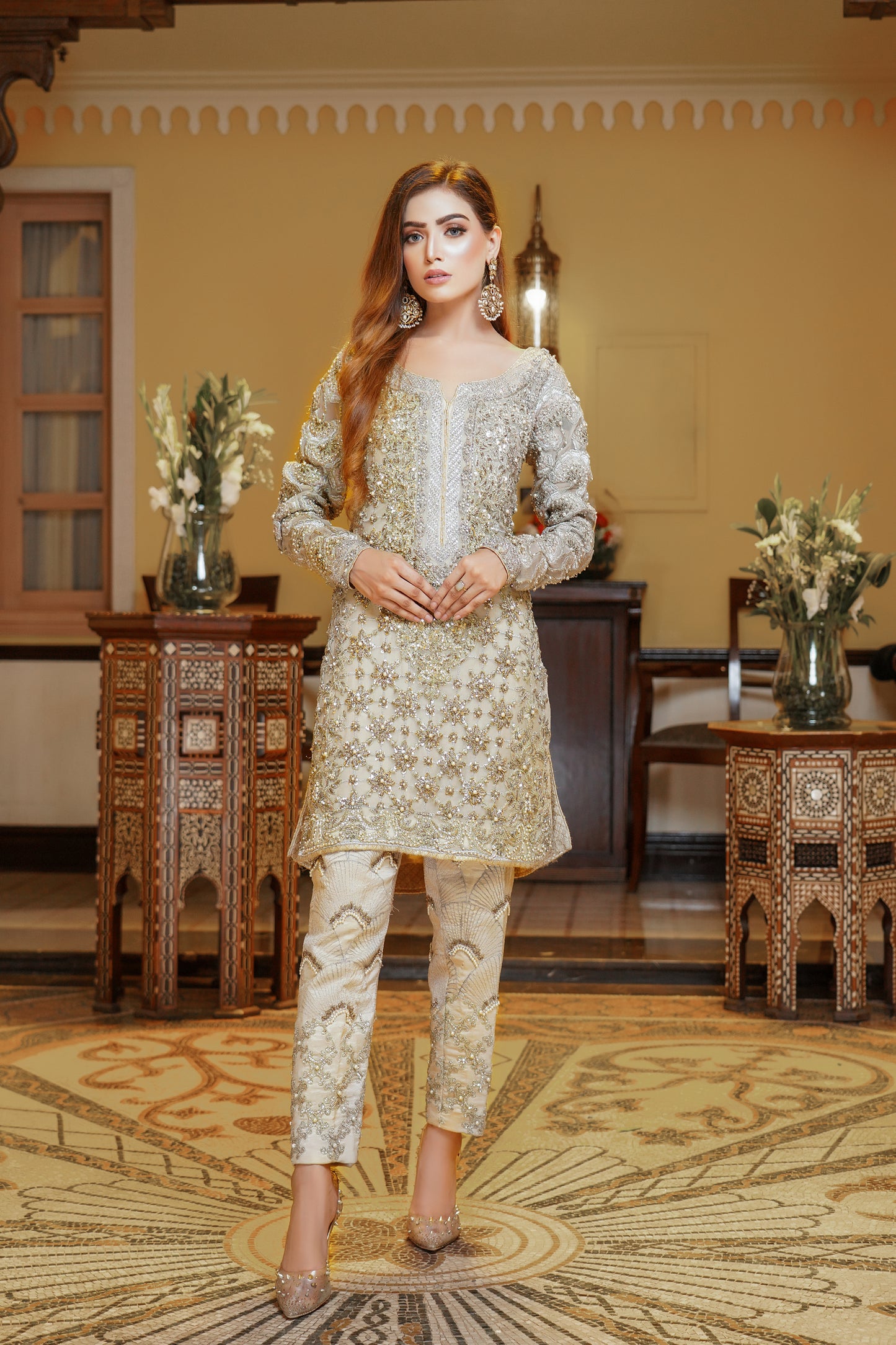 A light gold Pakistani formal dress with gold work and silver work around the neck. The dress has a fitted bodice, high neckline, and long sleeves, with intricate gold work detailing throughout the fabric. Silver work is present around the neckline, creating a beautiful frame for the wearer's face. The flowing skirt adds elegance and grace to the overall look. This dress is perfect for formal occasions and showcases the opulence and refinement of Pakistani fashion.