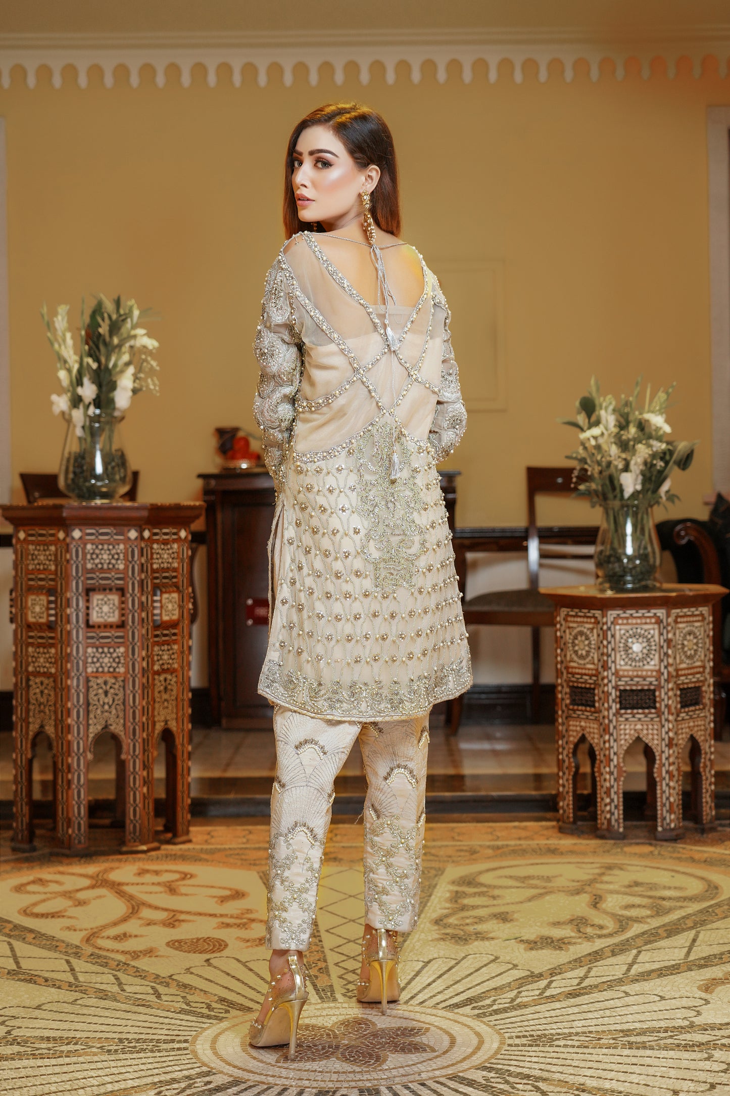 A light gold Pakistani formal dress with gold work and silver work around the neck. The dress has a fitted bodice, high neckline, and long sleeves, with intricate gold work detailing throughout the fabric. Silver work is present around the neckline, creating a beautiful frame for the wearer's face. The flowing skirt adds elegance and grace to the overall look. This dress is perfect for formal occasions and showcases the opulence and refinement of Pakistani fashion.