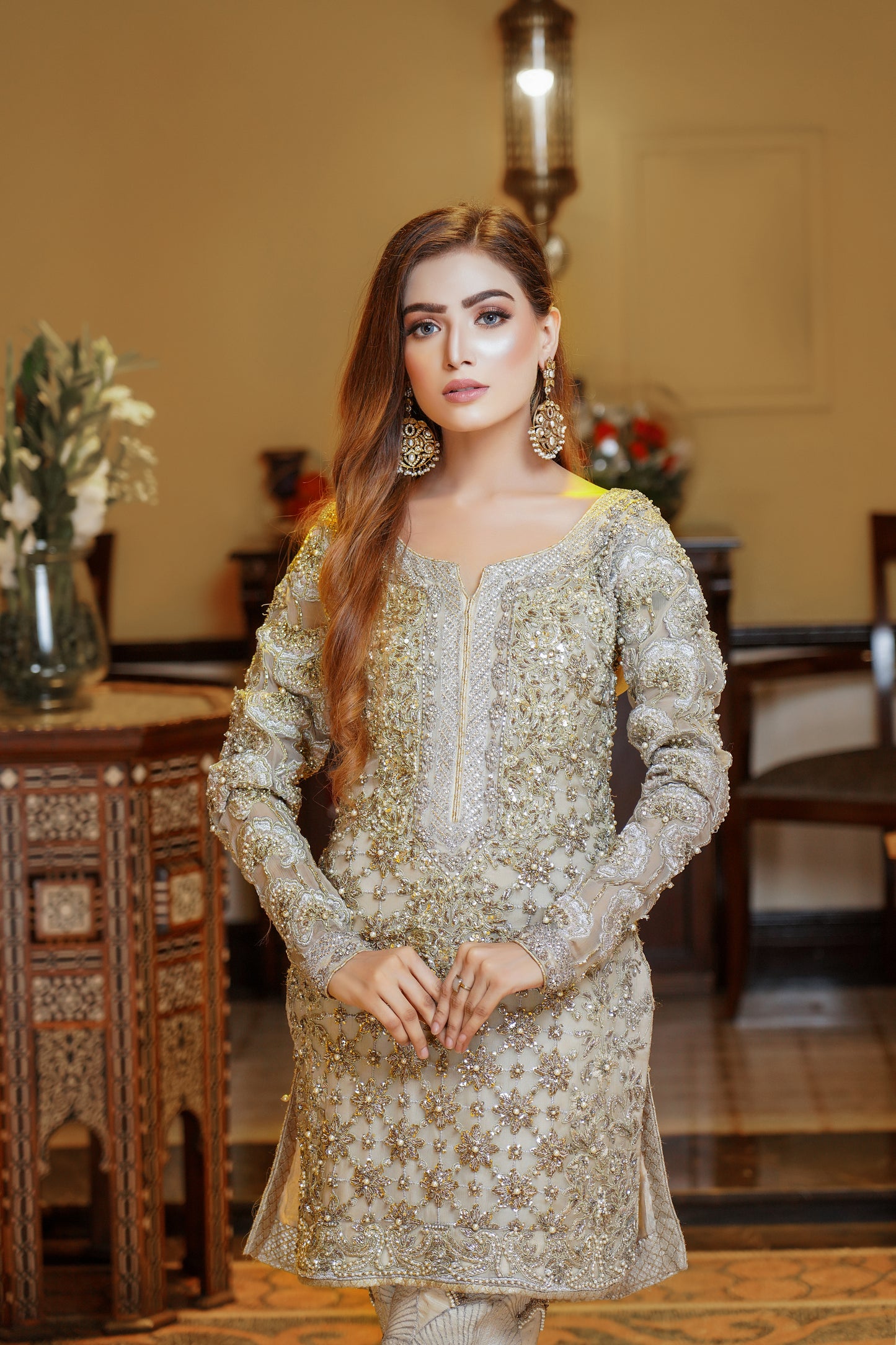 A light gold Pakistani formal dress with gold work and silver work around the neck. The dress has a fitted bodice, high neckline, and long sleeves, with intricate gold work detailing throughout the fabric. Silver work is present around the neckline, creating a beautiful frame for the wearer's face. The flowing skirt adds elegance and grace to the overall look. This dress is perfect for formal occasions and showcases the opulence and refinement of Pakistani fashion.