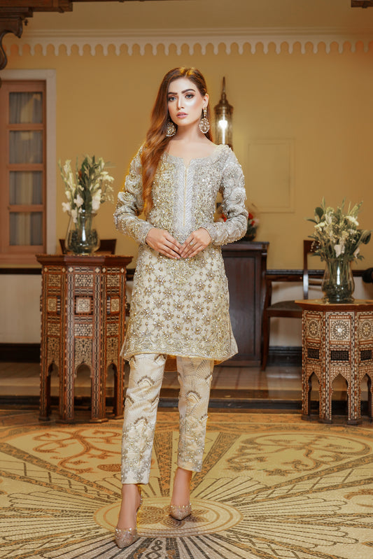 A light gold Pakistani formal dress with gold work and silver work around the neck. The dress has a fitted bodice, high neckline, and long sleeves, with intricate gold work detailing throughout the fabric. Silver work is present around the neckline, creating a beautiful frame for the wearer's face. The flowing skirt adds elegance and grace to the overall look. This dress is perfect for formal occasions and showcases the opulence and refinement of Pakistani fashion.