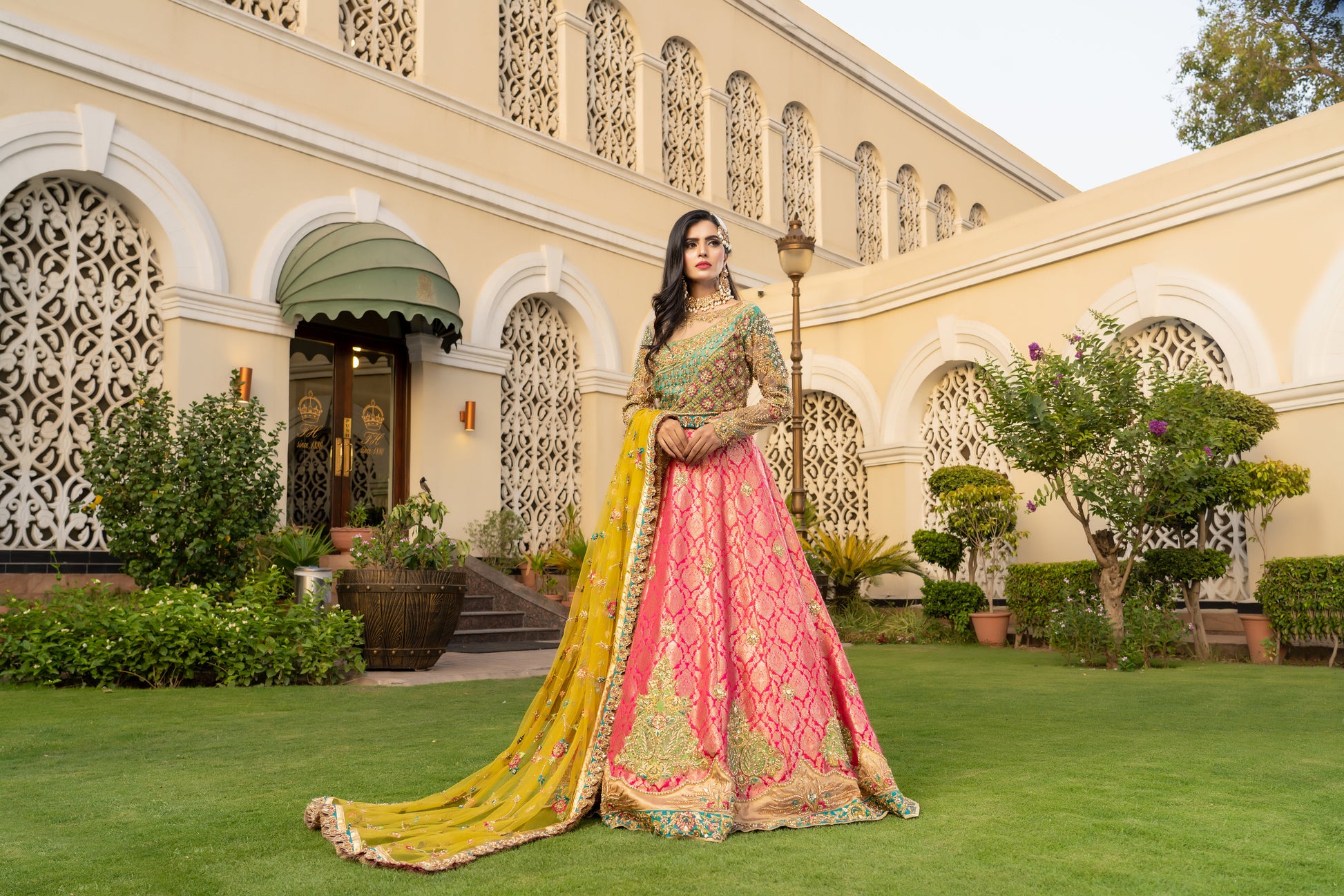 Pink and green Pakistani bridal dress with yellow dupatta. Intricate work embellishments, gold embroidery and beadwork on pink bodice, heavy border on green skirt. Flowing silhouette with fitted bodice and flared skirt. Vibrant yellow dupatta with delicate gold thread work. Perfect for Pakistani brides looking for colorful wedding outfits.