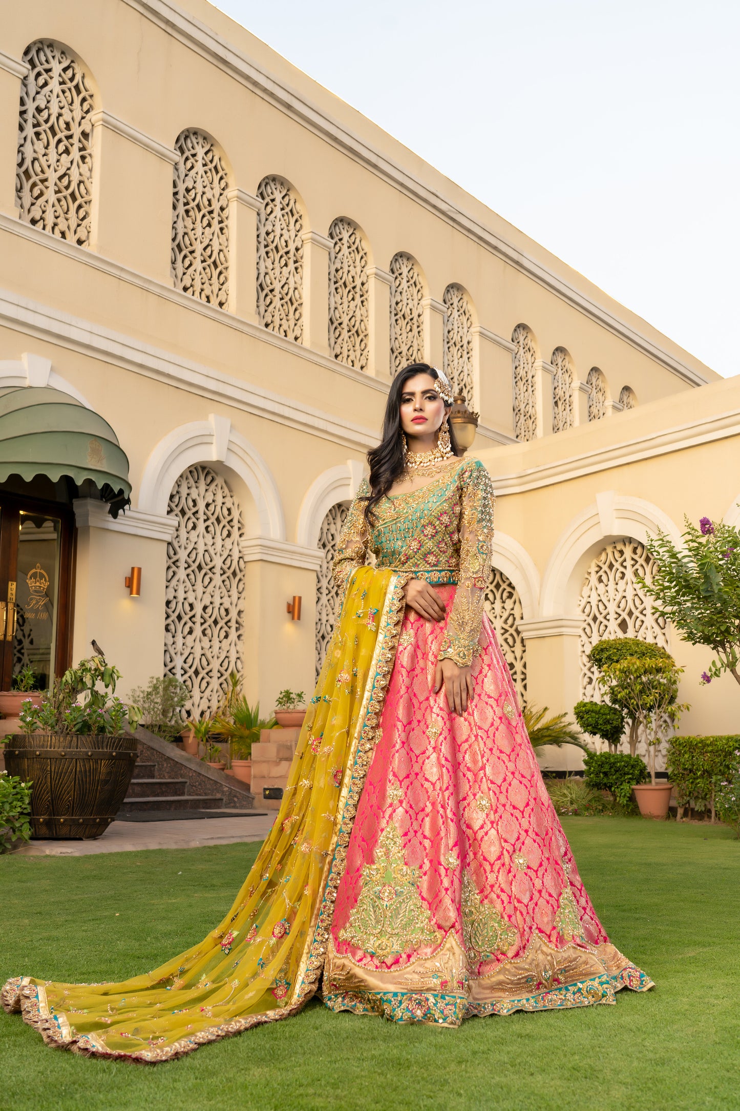 Pink and green Pakistani bridal dress with yellow dupatta. Intricate work embellishments, gold embroidery and beadwork on pink bodice, heavy border on green skirt. Flowing silhouette with fitted bodice and flared skirt. Vibrant yellow dupatta with delicate gold thread work. Perfect for Pakistani brides looking for colorful wedding outfits.