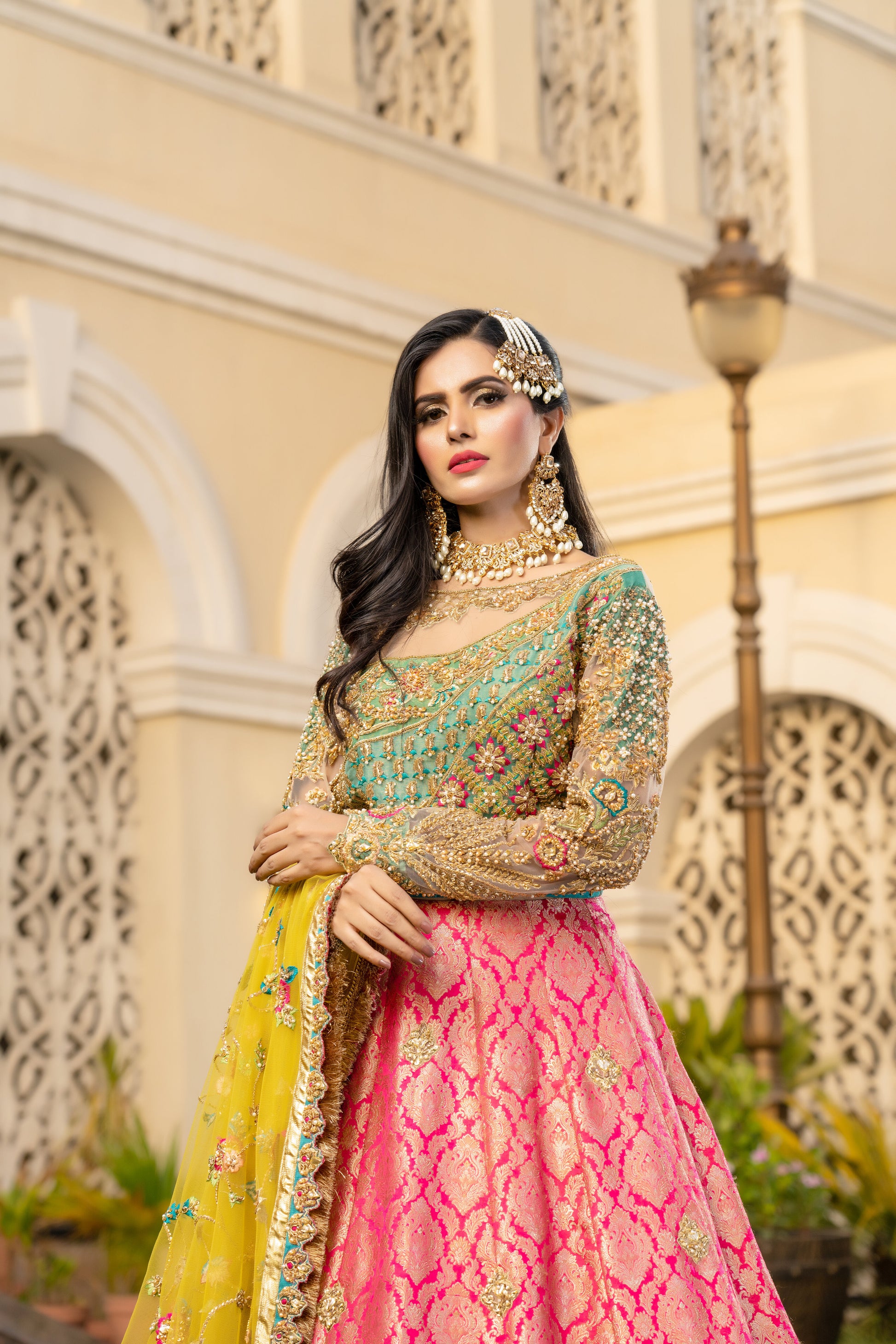 Pink and green Pakistani bridal dress with yellow dupatta. Intricate work embellishments, gold embroidery and beadwork on pink bodice, heavy border on green skirt. Flowing silhouette with fitted bodice and flared skirt. Vibrant yellow dupatta with delicate gold thread work. Perfect for Pakistani brides looking for colorful wedding outfits.