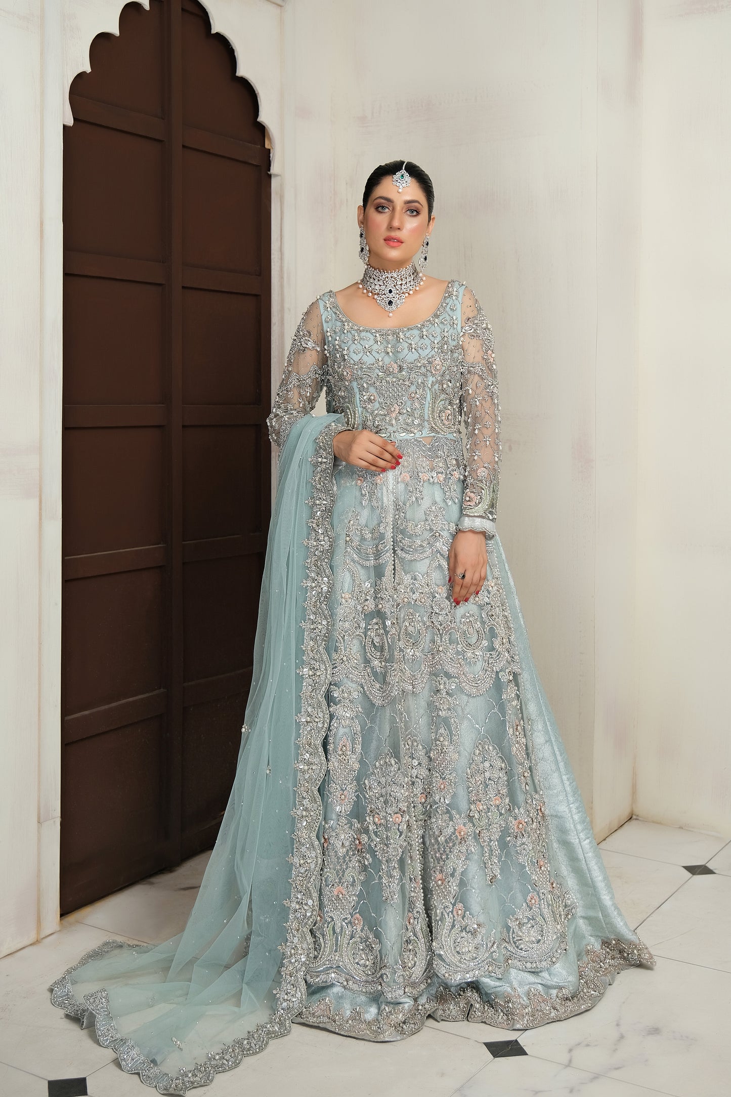 Pakistani ice blue bridal dress with silver work. The dress is a stunning shade of ice blue with intricate silver work embellishments. The neckline and sleeves are adorned with silver embroidery, and the skirt features a heavy silver border. The dress has a flowing silhouette with a fitted bodice and flared skirt. The outfit is completed with a matching dupatta that has a delicate silver border