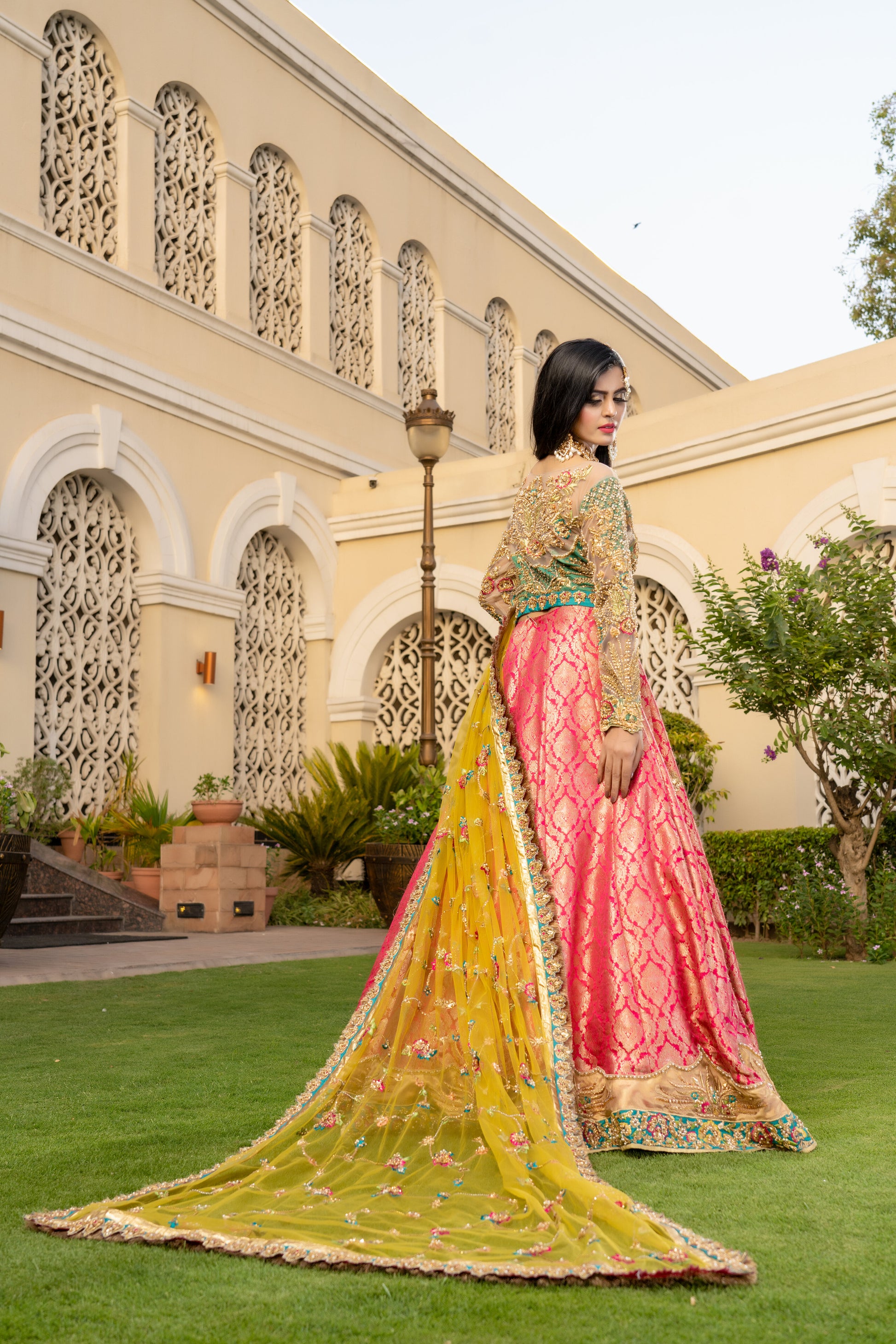 Pink and green Pakistani bridal dress with yellow dupatta. Intricate work embellishments, gold embroidery and beadwork on pink bodice, heavy border on green skirt. Flowing silhouette with fitted bodice and flared skirt. Vibrant yellow dupatta with delicate gold thread work. Perfect for Pakistani brides looking for colorful wedding outfits.