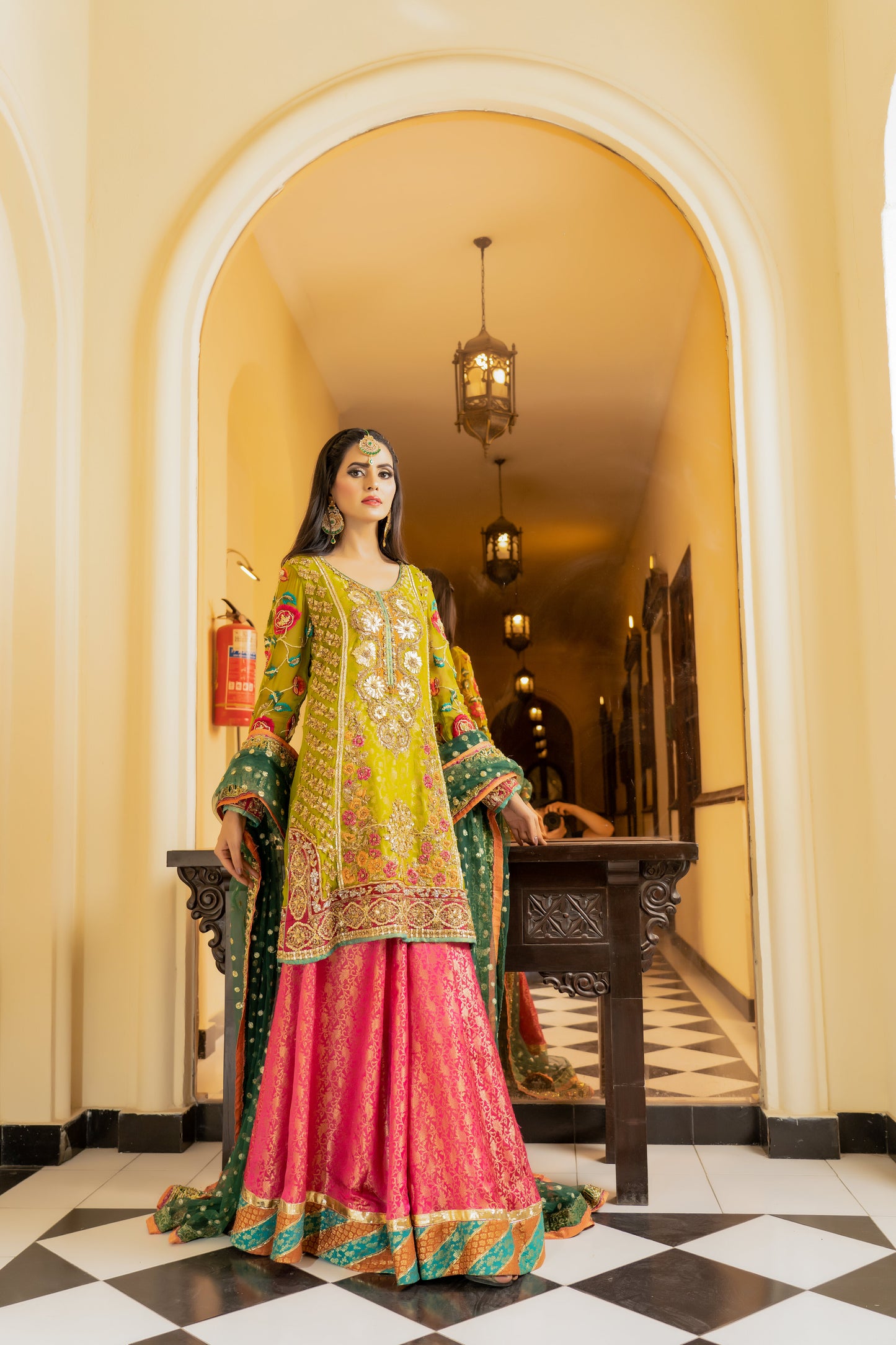 A stunning Pakistani bridal dress in yellow and pink with a green dupatta. Intricate gold handwork with embroidery, sequins, and beadwork in floral and paisley patterns. Fitted bodice with a sweetheart neckline and long sleeves, and a flowing skirt with a wide flare. The matching dupatta is draped over the head, adding a regal touch. Perfect for brides who want to make a bold and vibrant statement.