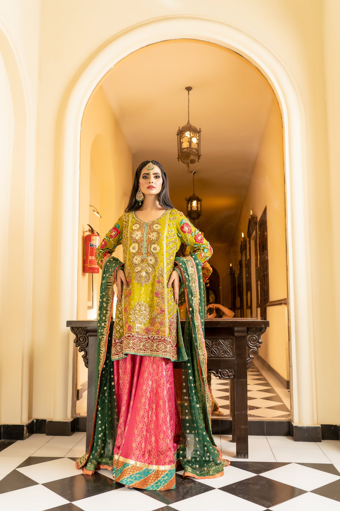 A stunning Pakistani bridal dress in yellow and pink with a green dupatta. Intricate gold handwork with embroidery, sequins, and beadwork in floral and paisley patterns. Fitted bodice with a sweetheart neckline and long sleeves, and a flowing skirt with a wide flare. The matching dupatta is draped over the head, adding a regal touch. Perfect for brides who want to make a bold and vibrant statement.