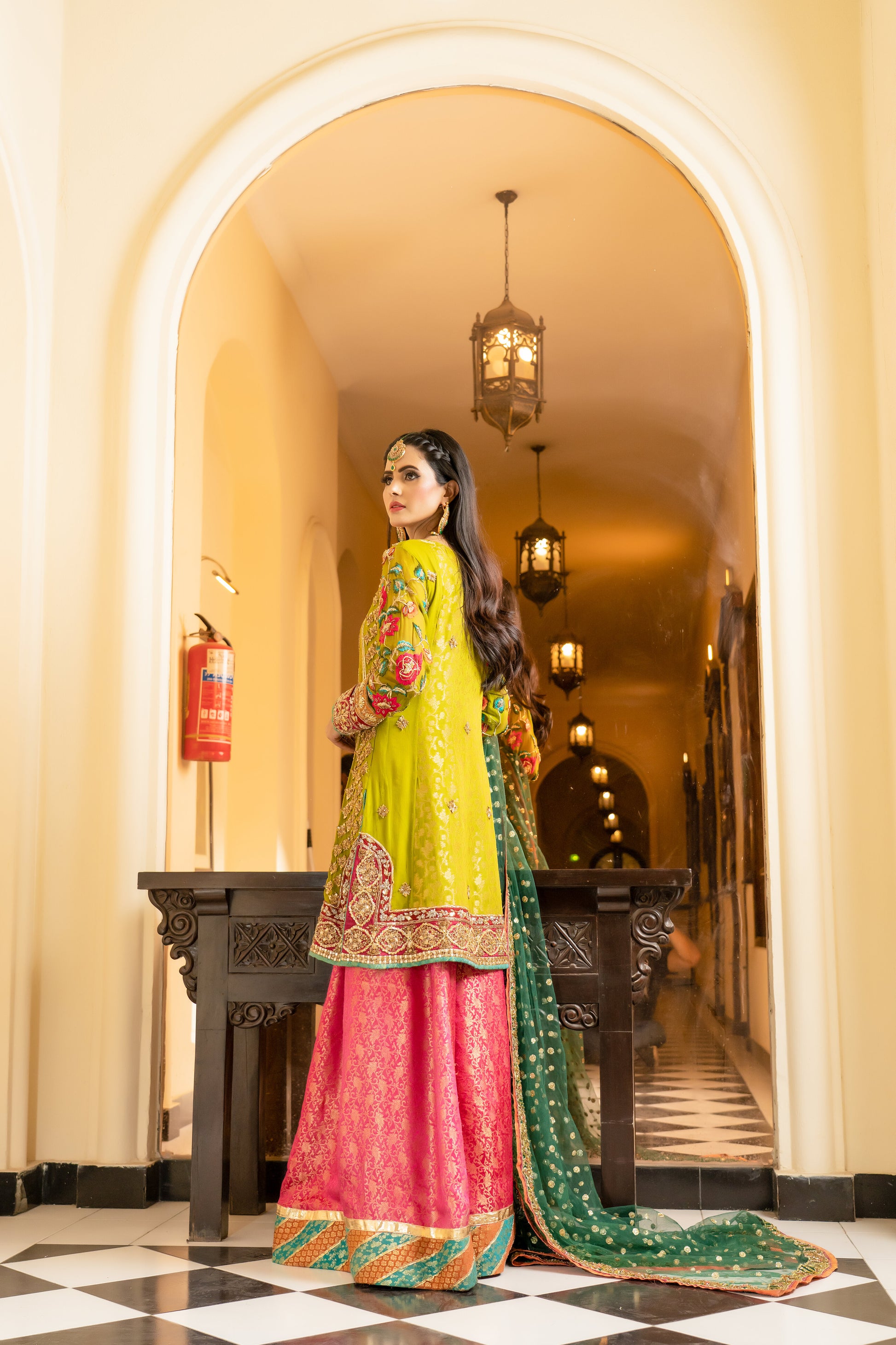 A stunning Pakistani bridal dress in yellow and pink with a green dupatta. Intricate gold handwork with embroidery, sequins, and beadwork in floral and paisley patterns. Fitted bodice with a sweetheart neckline and long sleeves, and a flowing skirt with a wide flare. The matching dupatta is draped over the head, adding a regal touch. Perfect for brides who want to make a bold and vibrant statement.