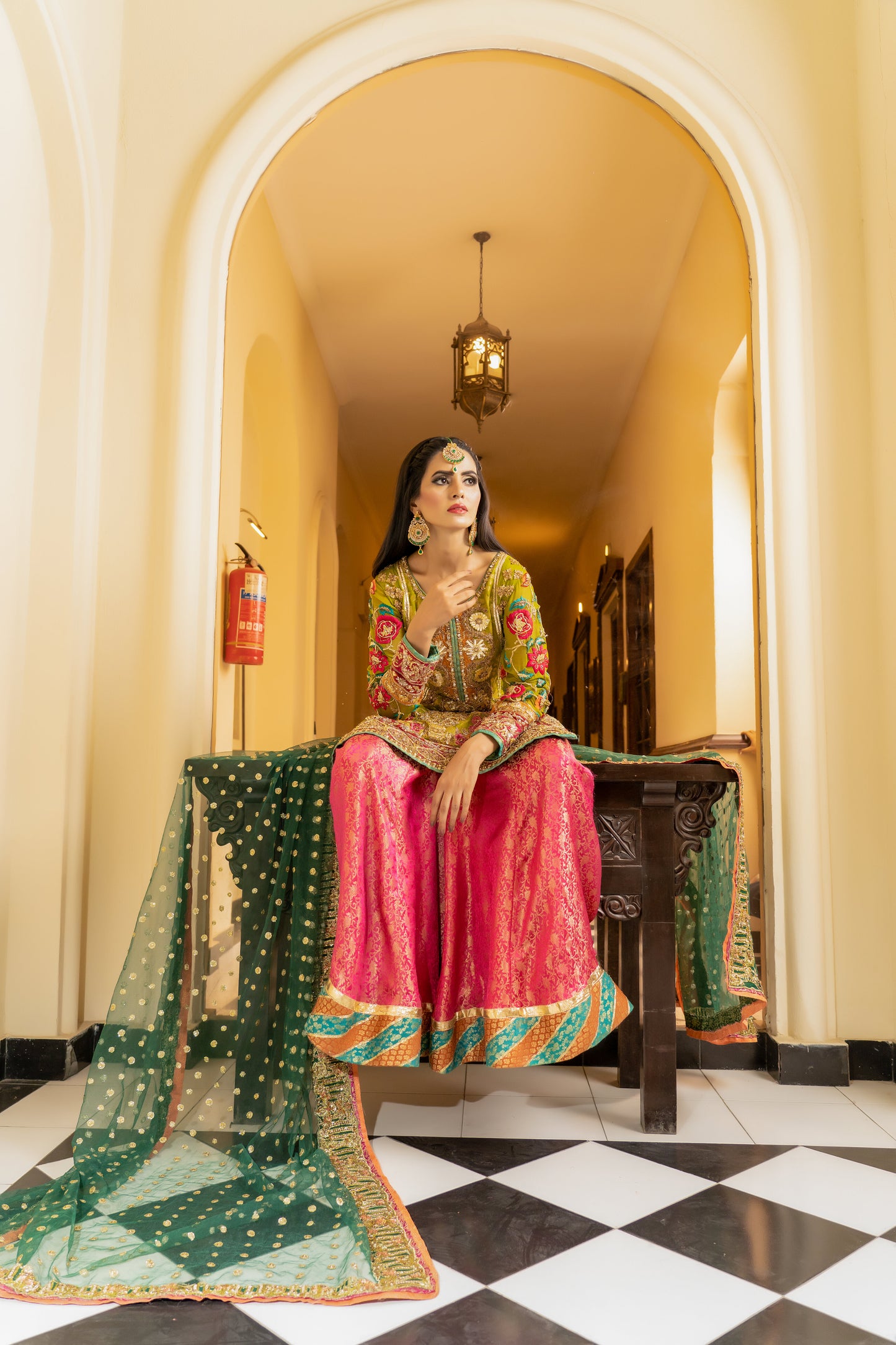 A stunning Pakistani bridal dress in yellow and pink with a green dupatta. Intricate gold handwork with embroidery, sequins, and beadwork in floral and paisley patterns. Fitted bodice with a sweetheart neckline and long sleeves, and a flowing skirt with a wide flare. The matching dupatta is draped over the head, adding a regal touch. Perfect for brides who want to make a bold and vibrant statement.