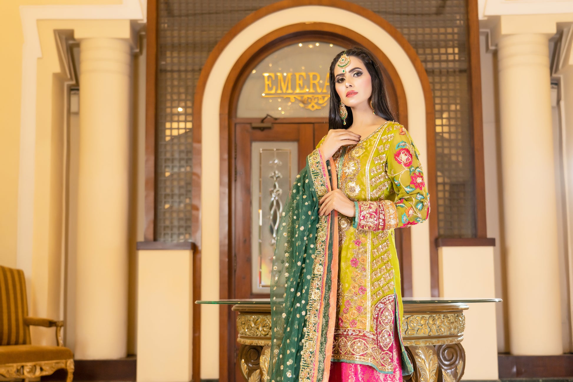 A stunning Pakistani bridal dress in yellow and pink with a green dupatta. Intricate gold handwork with embroidery, sequins, and beadwork in floral and paisley patterns. Fitted bodice with a sweetheart neckline and long sleeves, and a flowing skirt with a wide flare. The matching dupatta is draped over the head, adding a regal touch. Perfect for brides who want to make a bold and vibrant statement.