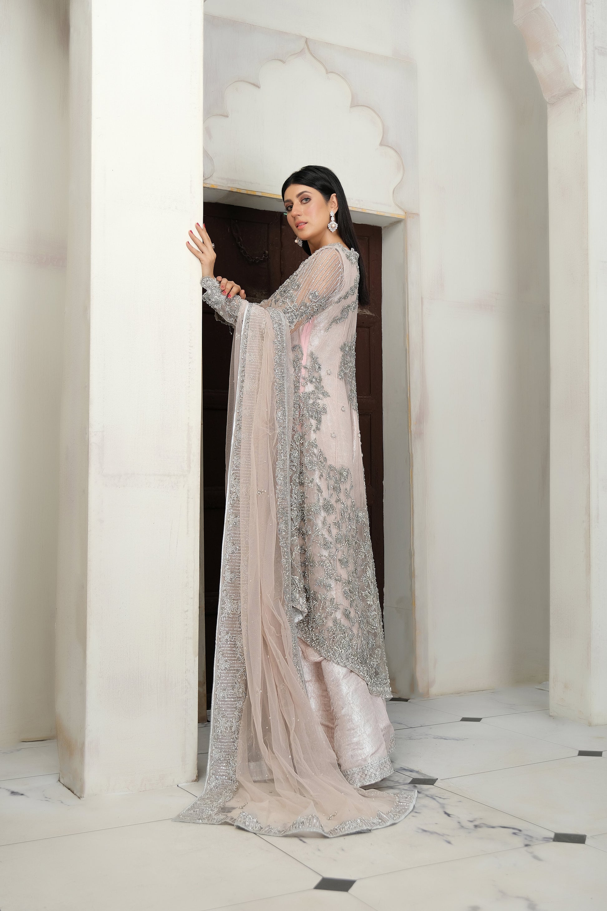 A beautiful blend of soft pink fabric and intricate silver embroidery, creating a striking contrast. The dress is elegantly adorned with silver details that flow seamlessly down the length of the dress, with a matching dupatta that adds an extra layer of grace. Perfect for any formal occasion, this dress is sure to impress with its delicate and eye catching design