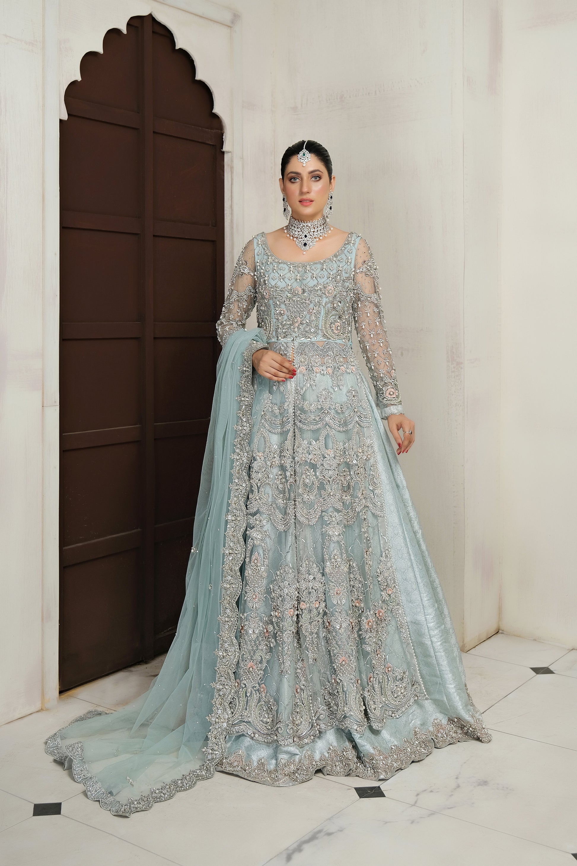 Pakistani ice blue bridal dress with silver work. The dress is a stunning shade of ice blue with intricate silver work embellishments. The neckline and sleeves are adorned with silver embroidery, and the skirt features a heavy silver border. The dress has a flowing silhouette with a fitted bodice and flared skirt. The outfit is completed with a matching dupatta that has a delicate silver border